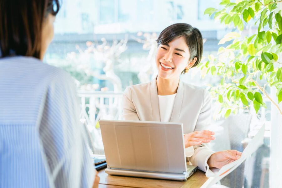明るい日差しが差し込む窓辺のオフィスで、笑顔で顧客と話すビジネスウーマン。彼女はライトベージュのジャケットを着用し、目の前のノートパソコンを挟んで書類を持ち、相手に説明しているようです。顧客の背中が写っており、活発なビジネスミーティングの様子がうかがえます。背景には緑の観葉植物が配置されています。