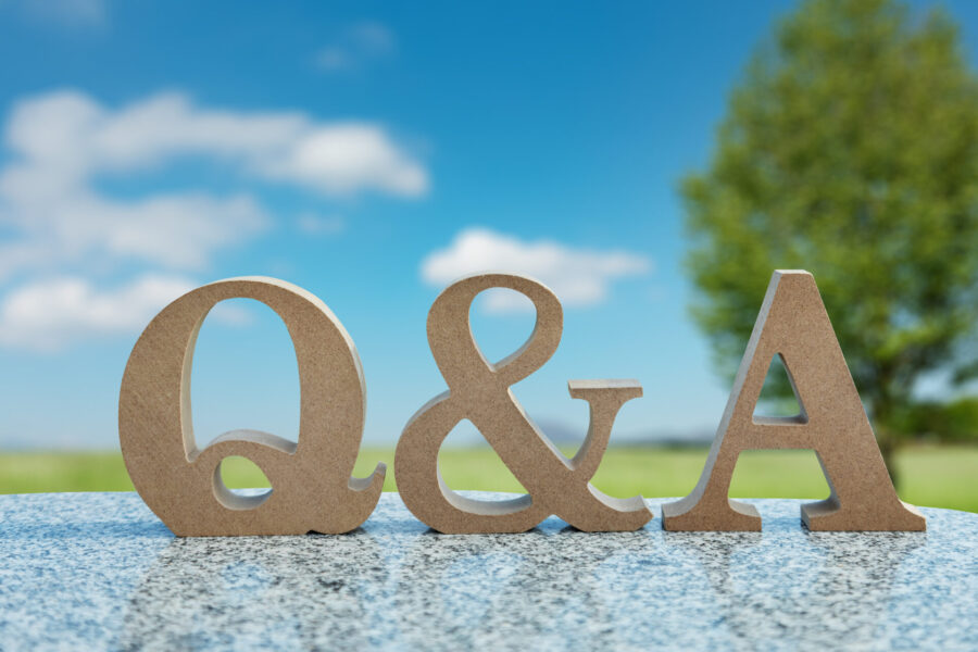 屋外の明るい背景に、木製の「Q & A」の文字がテーブルの上に立っている。青い空と緑の木々がぼやけて写っており、質問と回答、または疑問解決のコンセプトを表している。